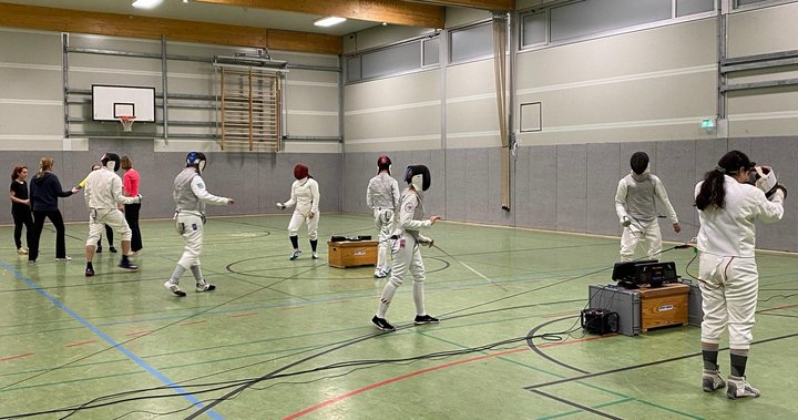 Ein Foto unseres Fechttrainings in einer Turnhalle. Im Vordergrund sind mehrere Degen- und Florett-Fechter*innen. Sie tragen jeweils vollständige weiße Fechtausrüstung , die Florett-Fechter*innen erkennt man an den silbernen E-Westen. Die Fechter*innen sind an Fecht-Meldegeräten angeschlossen und bereiten sich vor auf ihre Gefechte vor bzw. fechten. Im Hintergrund findet ein Anfänger*innen-Kurs statt. Die Teilnehmer*innen des Kurses tragen normale Sportkleidung. Die Halle hat den üblichen hellgrünen Turnhallenboden, die Wände sind grau, ganz hinten sieht man einen Basketballkorb und eine außer Reichweite in die Höhe gezogene Sprossenwand.